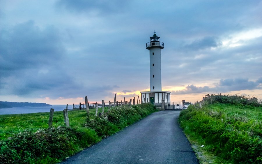 Sunset colours in the sky and a road that goes to a Lighthouse in Asturias, green grass on both sides of the road and the coast in the background